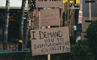 Photo showing series of placards in a stack in what appears to be a square or plaza. The bottom one reads 'I demand you to institutionalise equality'